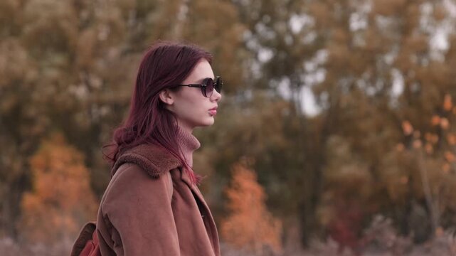 Teen girl wearing sunglasses and a brown coat, standing outdoors in autumn, looking serene against blurred fall foliage.