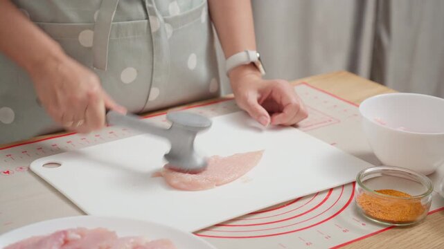 A cook tenderizes raw chicken breast with a kitchen hammer on a cutting board. Preparing fresh poultry meat for a healthy homemade recipe with spices for a protein-rich diet.