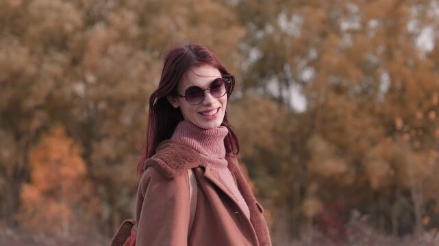 Teen girl standing with her back to the camera, turning around with a genuine smile, wearing sunglasses and a warm brown coat in a colorful autumn landscape.
