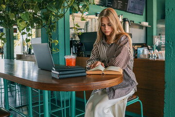 Smiling young woman sitting at cafe table with laptop, books and iced drink, reading a book in modern green interior