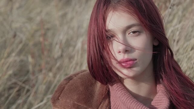 Close-up of teen girl with red hair and pink sweater, looking thoughtfully at the camera as wind blows strands of hair across her face in an autumn setting.