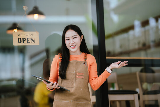 Smiling female barista in apron standing at café entrance, giving thumbs up, welcoming customers with joy and confidence.