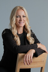 Portrait of a happy business woman wearing black suit sitting on the chair isolated on grey background, looking at the camera and smiling.