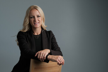 Portrait of a happy business woman wearing black suit sitting on the chair isolated on grey background.
