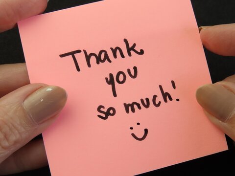 Woman hands holding pink note with the expression "Thank you so much" and a smile on black background. Thank you card.
