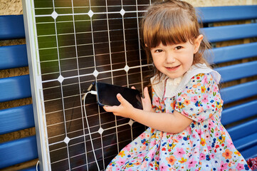 Young girl holds smartphone connected to solar panel for charging. Integration of sustainable...