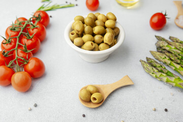 Green olives in ceramic bowl with tomatoes asparagus and seasonings on white table
