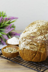 A loaf of bread rests on a wire rack next to a bowl of flour and another of seeds. The bread is whole wheat and has a crunchy texture. The simple scene has a sprig of lavender in the background.