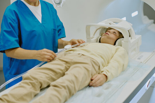 Caucasian child lying on MRI scanner bed undergoing medical imaging procedure while female healthcare worker in scrubs preparing equipment and monitoring patient during examination