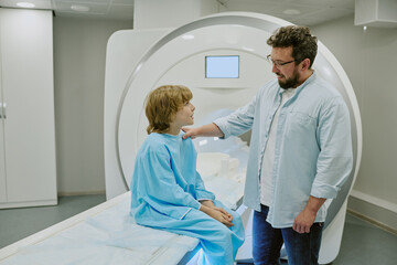 Caucasian middle aged man comforting Caucasian child sitting on MRI scanner in medical facility, child wearing hospital gown, man gently touching child's shoulder, both interacting before procedure