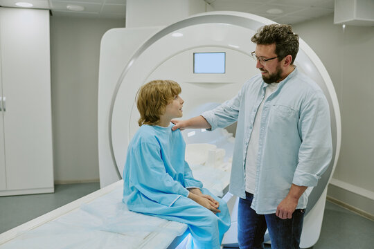 Caucasian middle aged man comforting Caucasian child sitting on MRI scanner in medical facility, child wearing hospital gown, man gently touching child's shoulder, both interacting before procedure
