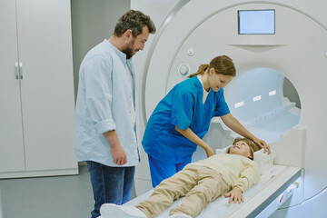 Female medical professional preparing Caucasian child for MRI scan while middle aged Caucasian man standing nearby observing procedure in modern hospital imaging room