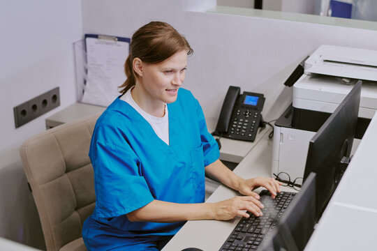 Caucasian middle aged woman wearing medical scrubs working at computer in modern office setting, sitting at desk and typing on keyboard with printer and phone visible in background