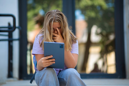 Upset schoolgirl sitting on stairs and covering face while holding tablet.  
