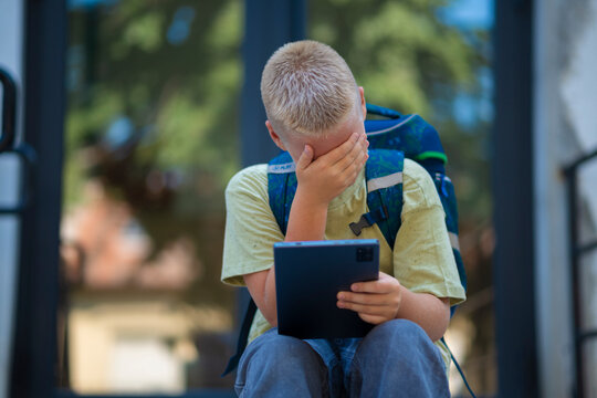 Sad schoolboy with backpack sitting on stairs and covering face while holding tablet.  
