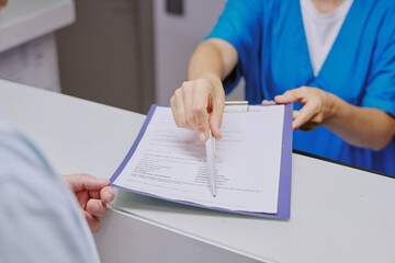 Caucasian middle aged woman handing clipboard with medical form and pen to Caucasian middle aged man at reception desk, both partially visible, focus on hands and document