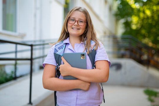 Smiling schoolgirl with backpack holding digital tablet in front of school.  
