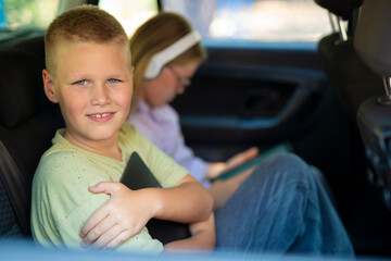 Smiling schoolboy holding tablet while sitting in car with his sister on the way to school.  
