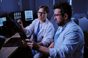 Caucasian young adult woman and Caucasian young adult man wearing medical coats analyzing brain scan images together in hospital radiology room, with computer monitors displaying MRI results