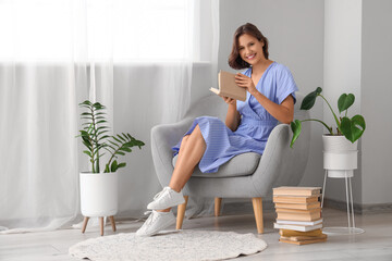Pretty young woman reading book on armchair at home