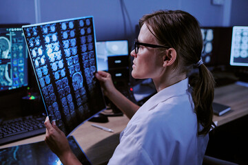 Caucasian middle aged woman wearing glasses analyzing brain MRI scans in medical office, sitting at desk with multiple computer monitors displaying diagnostic images