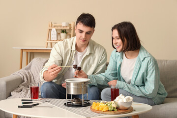 Young happy couple with glasses of wine dipping marshmallows into chocolate fondue at home