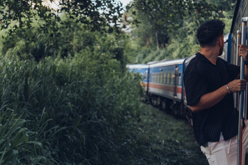 Fototapeta premium Young man holding handrail while traveling by train through lush tropical vegetation in Sri Lanka