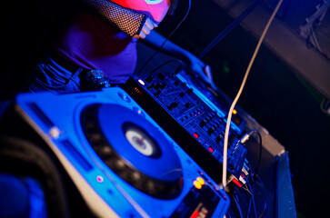 DJ's hand deftly manipulates turntable and mixer bathed in blue and red lights within nightclub. Vibrant club lighting highlights equipment and DJ wearing headset for cueing tracks, capturing essence