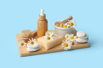 Wooden tray with chamomile flowers, soap bar and spa stones on blue background, closeup