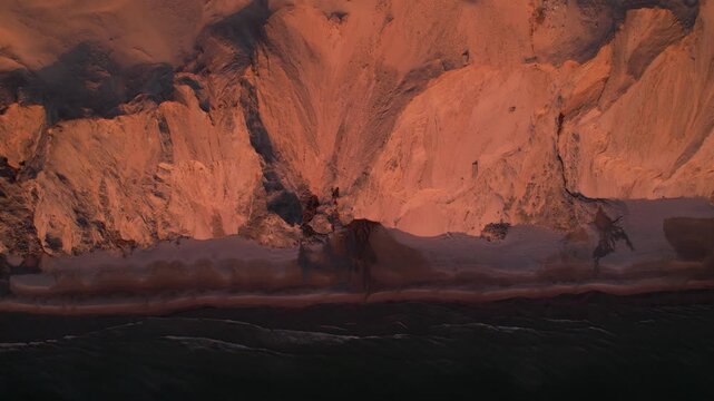 Aerial top view of dramatic coastal sand cliffs and shoreline at sunset. Natural erosion patterns with orange evening light reflecting on the beach and sea in Northern Denmark