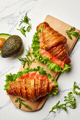 Delicious croissant sandwiches with salmon, lettuce, and avocado on wooden board
