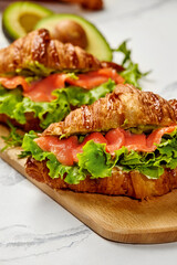 Delicious croissant sandwiches with salmon, avocado, and lettuce on wooden board