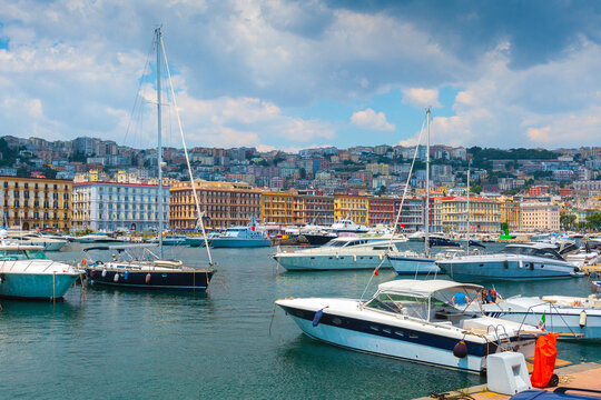 Yachts and sailboats are moored in the Molo Sannazzaro marina in Naples, Italy. The dense, colorful cityscape rises on the hill behind the busy harbor. - Powered by Adobe