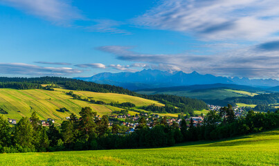 Picturesque village set among golden-green fields and dark pine forests, with dramatic mountain peaks rising in the background under a vivid blue sky.
