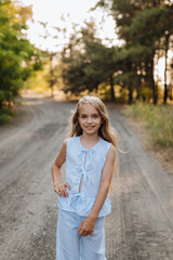 Fototapeta premium Beautiful nine-year-old blonde girl with long hair in a blue suit, walking in nature in the forest. Summer, sunset