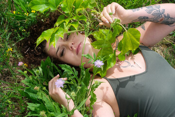 A tattooed woman lies in tall grass, her face partly hidden behind green leaves and wildflowers. A sensual, natural summer portrait evoking freedom, femininity, and connection to nature