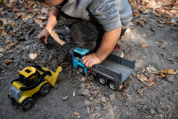 Fototapeta premium Toddler playing with toy trucks in the dirt and leaves