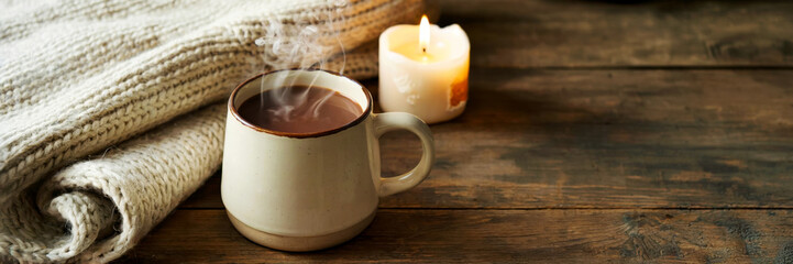 Rustic wooden table with steaming coffee in cream mug. Beige knitted blanket and lit candle create warm and cozy feeling. Soft natural light and negative space enhance calm mood, concept of relaxation