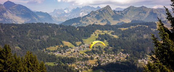 Flying paraglider passing by in stunning French Alps scenery with Mont Blanc massive in the background and Les Gets town in valley below. Anamorphic cinematic panorama.