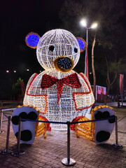 A large New Year's light installation in the form of a teddy bear, decorated with garlands and a bright illumination of many colorful light bulbs. A bear cub in a red bow, urban on the square.