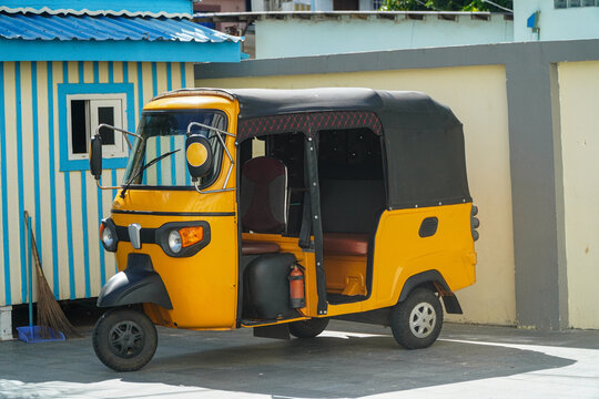 A vibrant yellow auto rickshaw contrasts with a striped building, embodying culture and colorful simplicity. Its open design and compact form captures transportation's essence and a tropical essence.