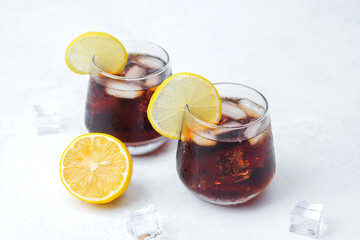Two glasses of dark soda with lemon slices and ice cubes served on light background with citrus garnish