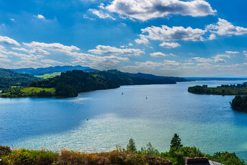 Wide panorama of Czorsztyn Lake with sailboats gliding across shimmering water, seen from the castle ruins. The green hills and dramatic sky frame this peaceful landscape.