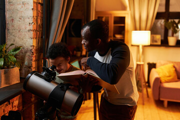 Obraz premium Mature African American father standing in front of telescope holding book telling his son about universe, when watching stars using telescope