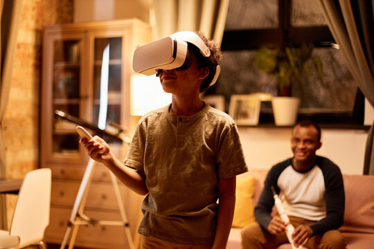 Medium portrait of joyful African American boy using VR headset to learn something, his father sitting on couch holding toy rocket looking at him