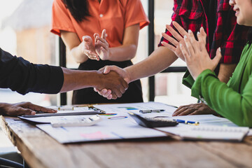 Business partners shaking hands at a meeting while their coworkers are clapping, celebrating the closing of a deal or a successful agreement.
