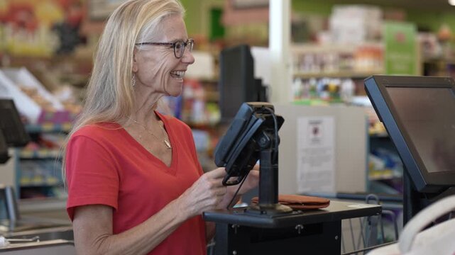 An attractive mature woman smiles while checking out her items at a grocery store. She is engaged and appears happy during her shopping trip.