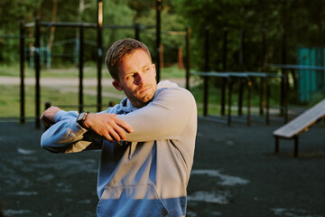 Caucasian young adult man stretching arms outdoors on sports ground, preparing for workout, looking away from camera, wearing smartwatch, surrounded by exercise equipment and trees