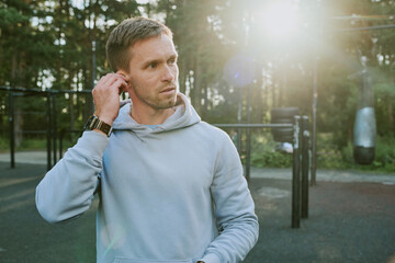 Caucasian young adult man adjusting wireless earbud while standing outdoors in park with fitness equipment, sunlight shining through trees in background, wearing casual hoodie