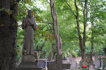 Fototapeta premium Stone statue of a monk holding a cross with a skull at the base, standing among cemetery graves and trees.